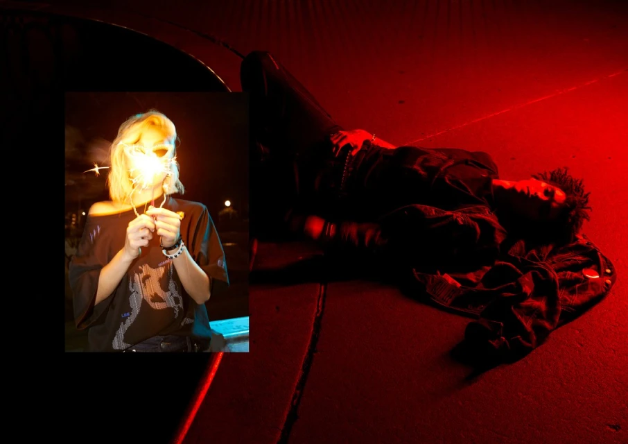 Two individuals, one lying on a red surface in dim lighting, the other holding sparklers in front of a dark background.