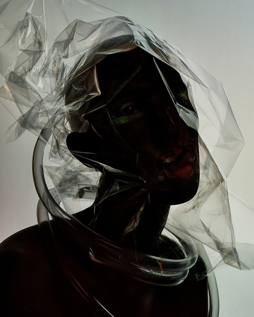 Dramatic fashion portrait: person enveloped in transparent plastic with clear tubes around the neck against a muted background.