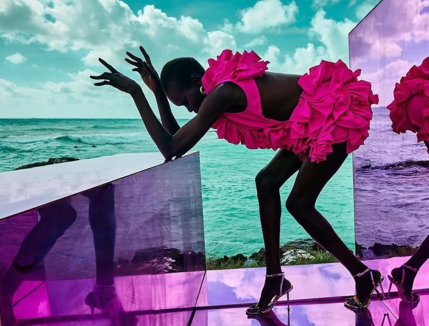 Two models in ruffled pink dresses pose on reflective cubes against an ocean backdrop, creating a surreal and vibrant fashion image.