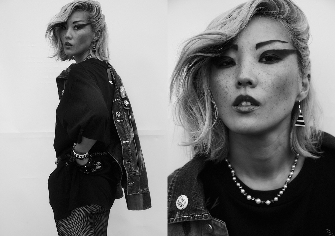Monochrome fashion portrait: Asian woman with dramatic eye makeup, denim jacket, jewelry, and platinum blonde hair against a plain background.