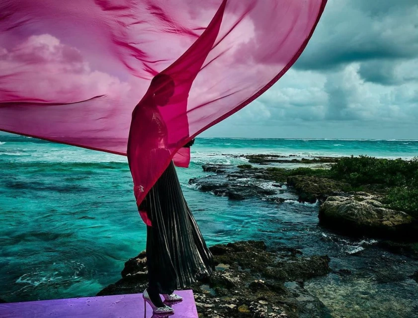 Woman on purple platform near ocean with flowing magenta fabric. Model wears silver stilettos, black skirt with ocean waves in background.