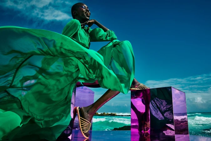 Black woman in a flowing green dress seated on a reflective purple cube against a bright ocean background by James Macari