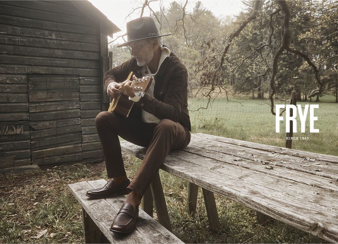 Adult man in hat playing acoustic guitar sitting on a wooden bench outside in a rural setting near a small wooden building.