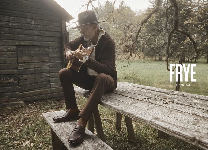 Adult man in hat playing acoustic guitar sitting on a wooden bench outside in a rural setting near a small wooden building.
