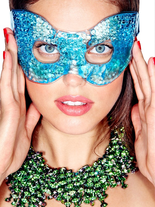 Woman wearing a blue beaded eye mask and green beaded necklace with red nail polish against a white background in a close-up shot.