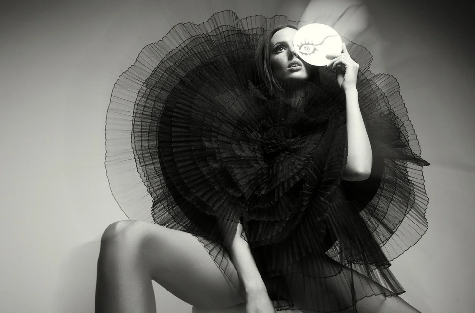 Monochrome portrait of a seated adult female in a ruffled dress, holding a round prop with a drawn eye, gazing upwards in a studio.