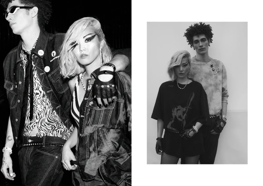 A black and white fashion-style photo featuring two young adult models in stylish attire, standing indoors with distinctive makeup and accessories.