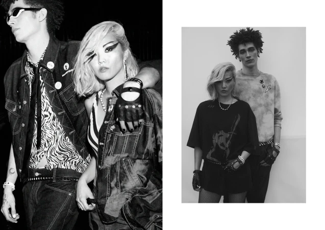 A black and white fashion-style photo featuring two young adult models in stylish attire, standing indoors with distinctive makeup and accessories.