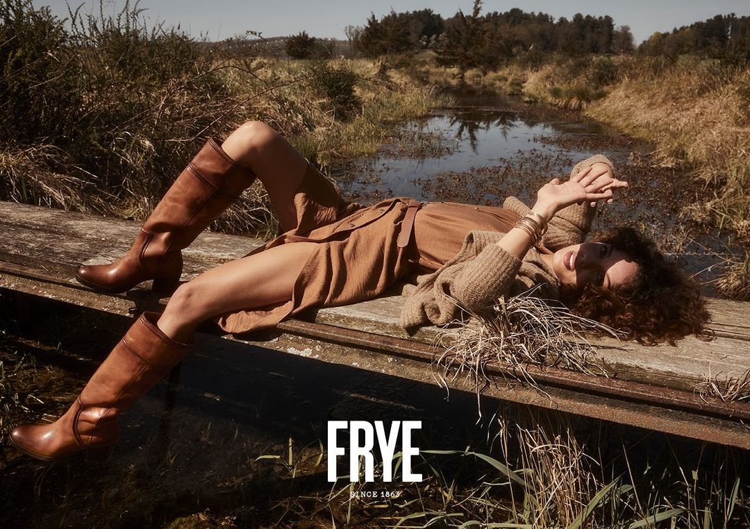 A woman in a brown dress and leather boots reclines on a wooden surface in a marshy field for a Frye fashion advertisement.