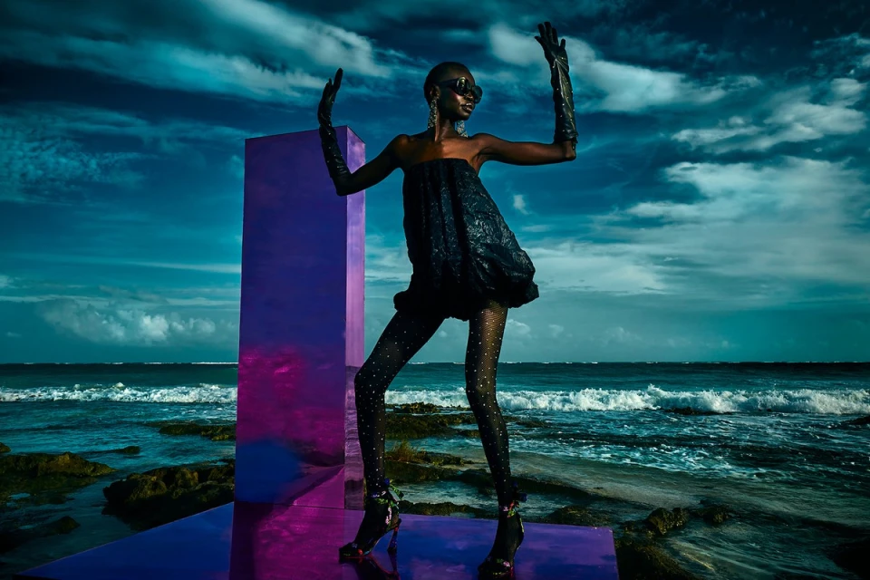 Fashionable woman in a black dress and floral heels striking a pose on a reflective platform with an ocean and blue sky background.