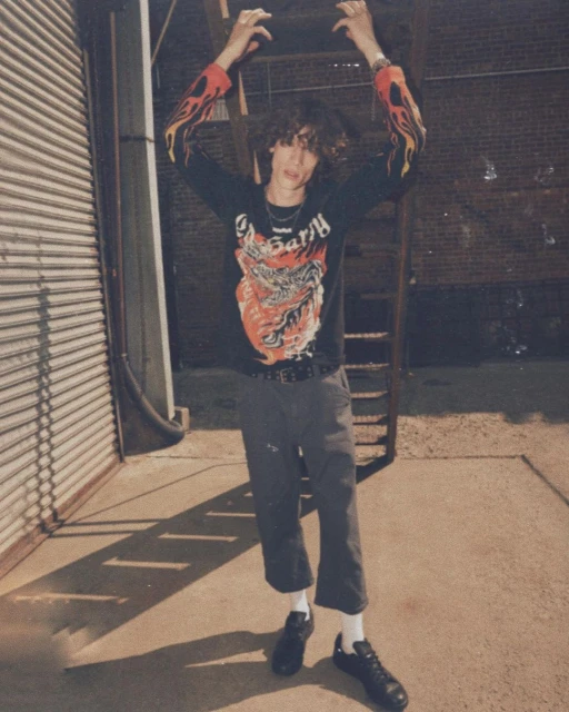 Adult male poses in an urban alleyway wearing a graphic long-sleeve shirt, dark pants, and black sneakers with a metal fire escape.