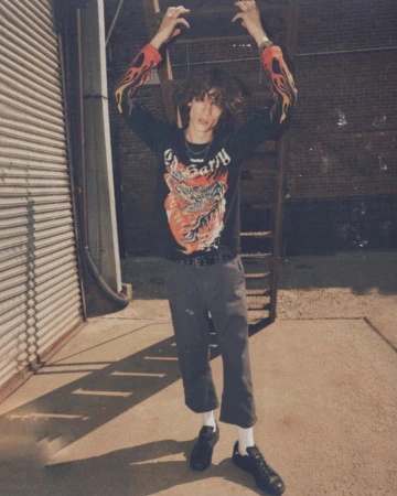 Adult male poses in an urban alleyway wearing a graphic long-sleeve shirt, dark pants, and black sneakers with a metal fire escape.