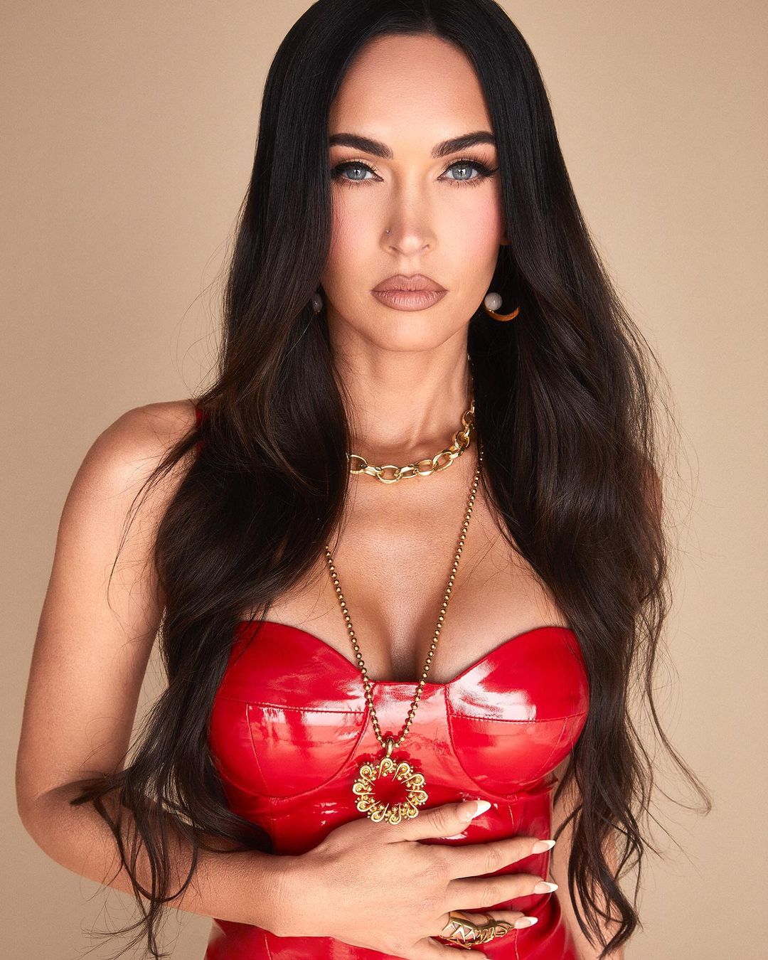 Portrait of a brunette woman with blue eyes wearing a red patent leather top and gold jewelry against a beige background.