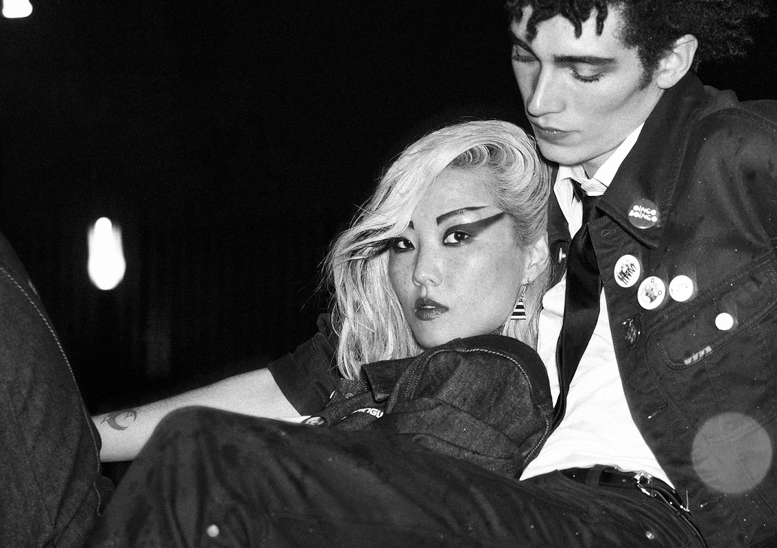 Monochrome portrait of an Asian woman with striking eye makeup alongside a Caucasian man with curly hair, both in denim jackets.