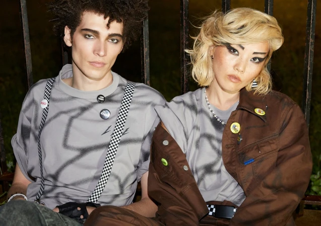 Two models with dramatic makeup posing outdoors against a dark iron fence, shot by James Macari.