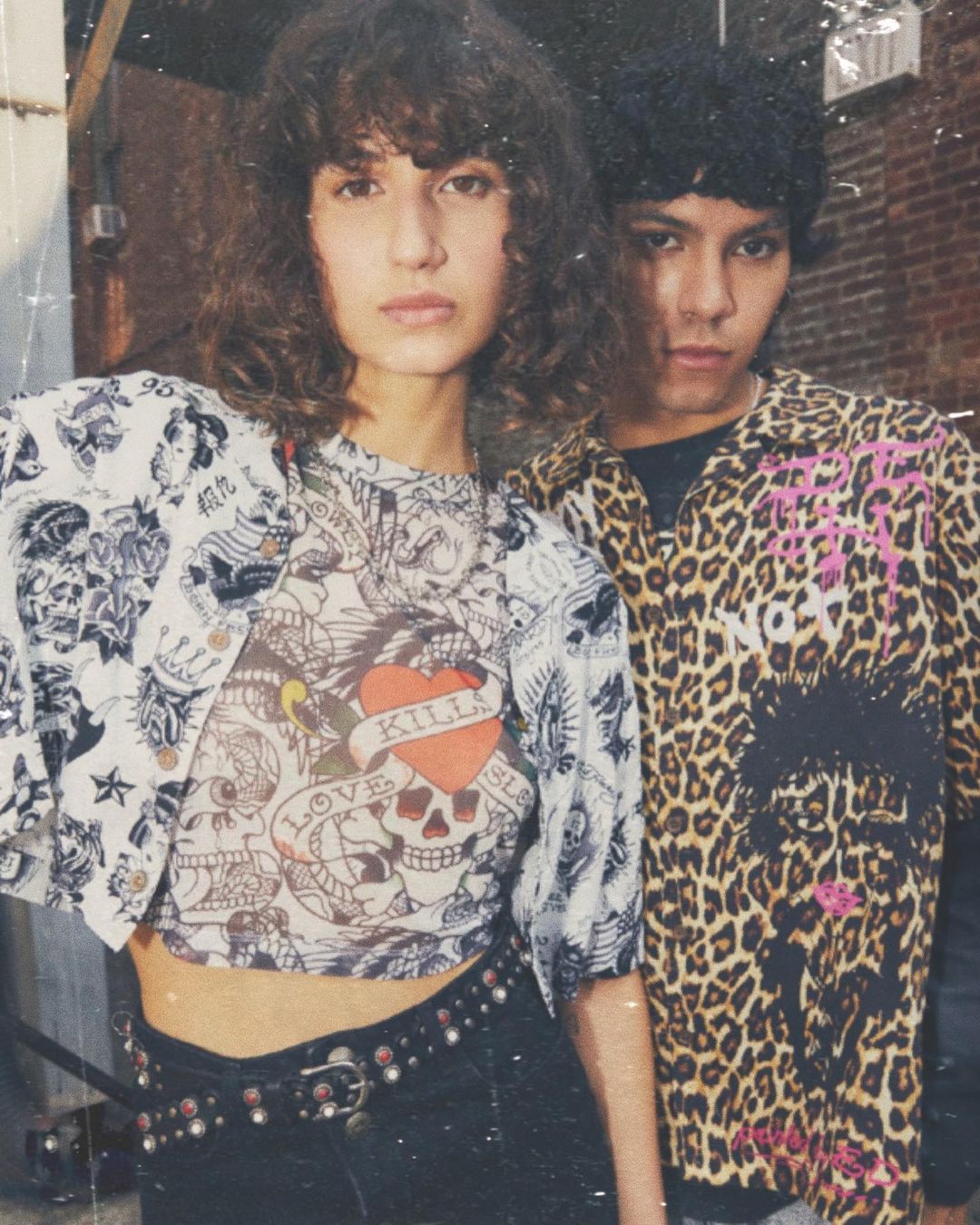 Two fashion models strike a pose outdoors. The woman wears a tattoo-style graphic crop top with black pants, while the man has a leopard print button-down.