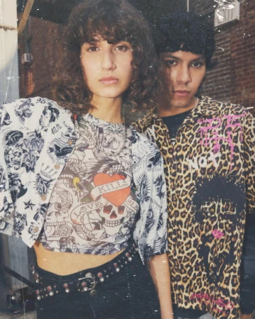 Two fashion models strike a pose outdoors. The woman wears a tattoo-style graphic crop top with black pants, while the man has a leopard print button-down.