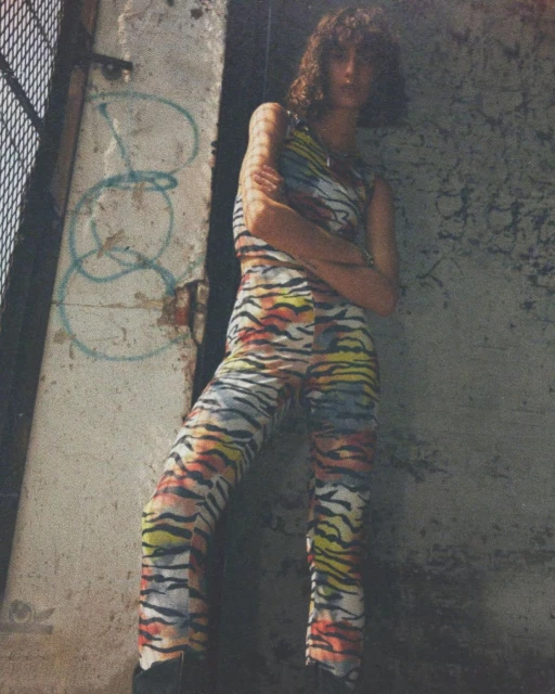 Model poses in colorful, patterned jumpsuit, standing against a weathered concrete wall with graffiti in an urban outdoor setting.