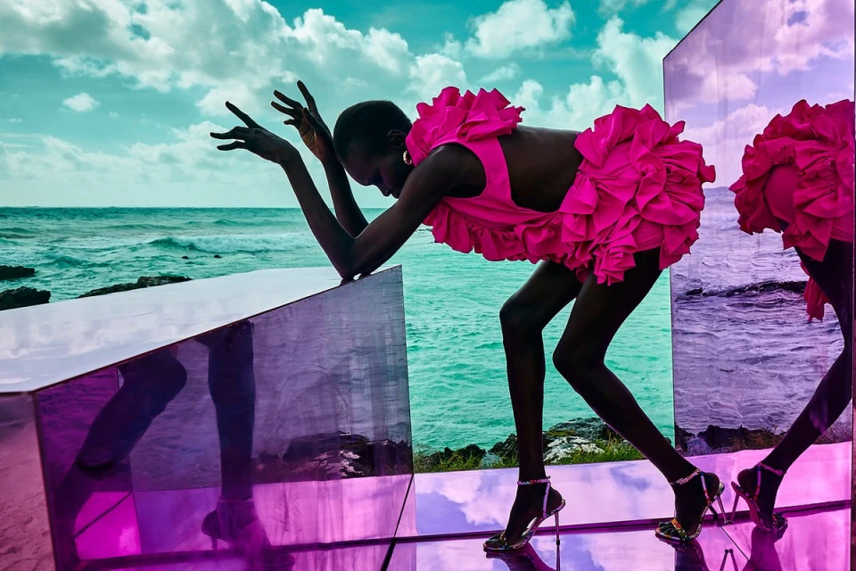 Woman in bright pink ruffled dress poses on mirrored cube at ocean. Model leans on display with ocean and sky background in fashion shot.
