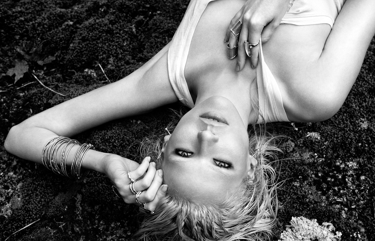 Black and white portrait of a blonde woman lying on moss, adorned with multiple rings and bracelets in an outdoor setting.