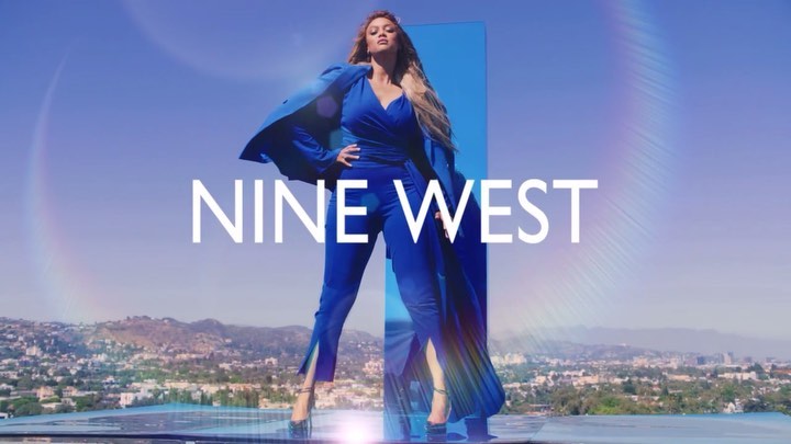An adult female fashion model poses in a blue pantsuit by a reflective surface with a mountain range and sky background, advertising Nine West.
