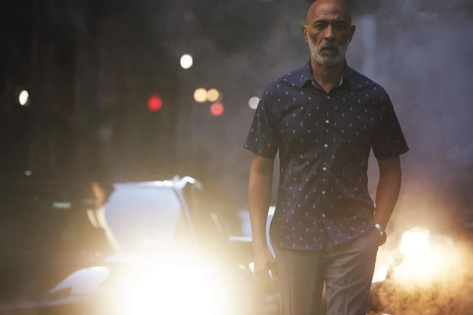 Adult male wearing a patterned shirt and slacks, walking in an outdoor environment with a car and streetlights visible in the background.