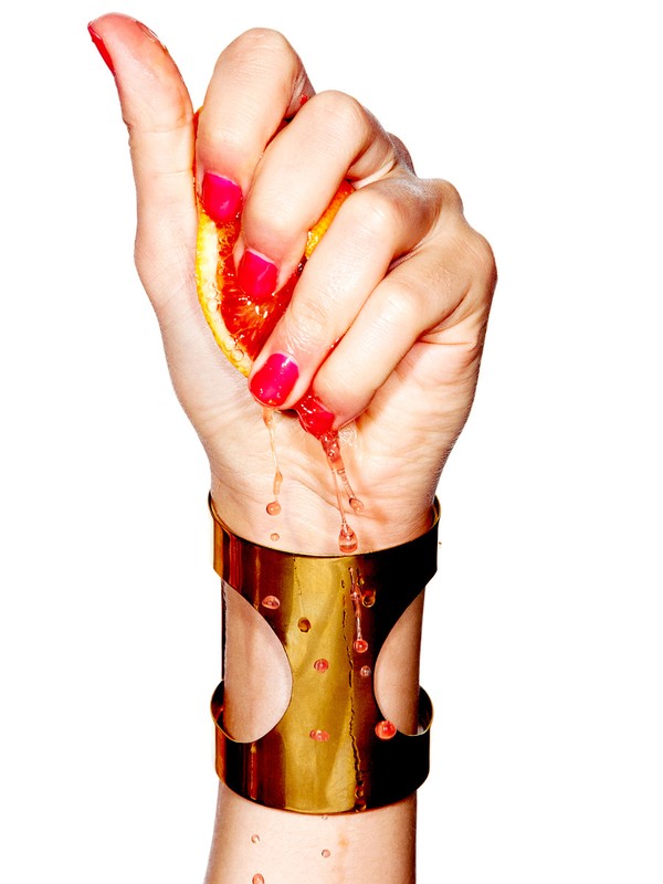 Close-up of a fair-skinned woman's hand squeezing half an orange, with orange juice dripping onto a gold cuff bracelet, isolated on white.