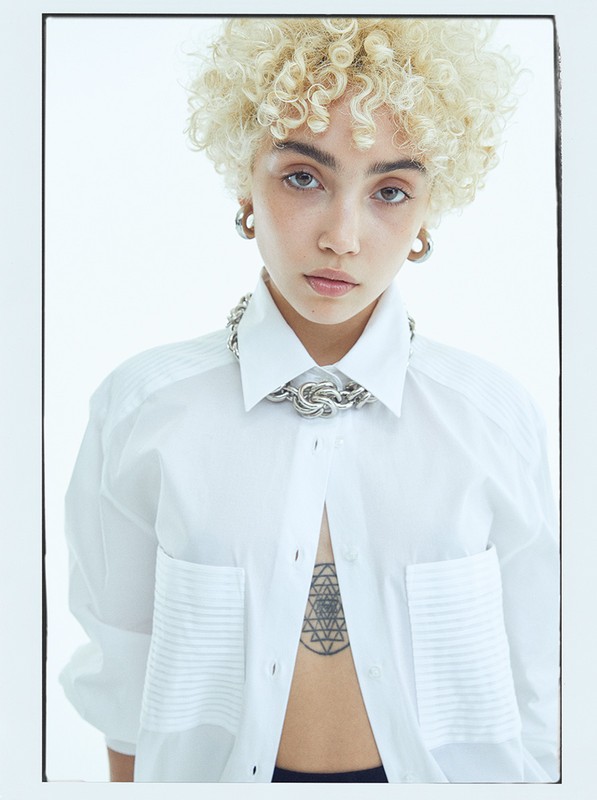A woman with curly blonde hair and a silver chain poses in a white shirt with a stern expression against a white backdrop.