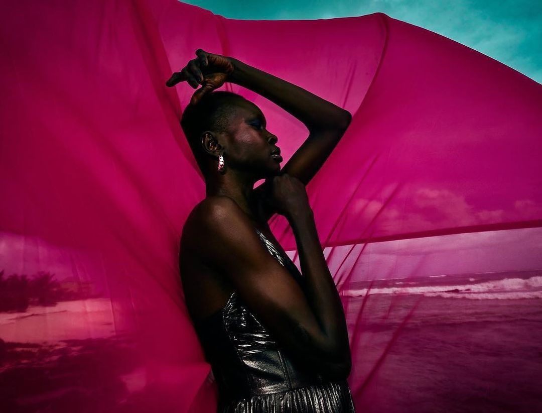Woman in shiny dress poses with pink fabric against ocean backdrop in a striking fashion shot with dramatic lighting and styling.
