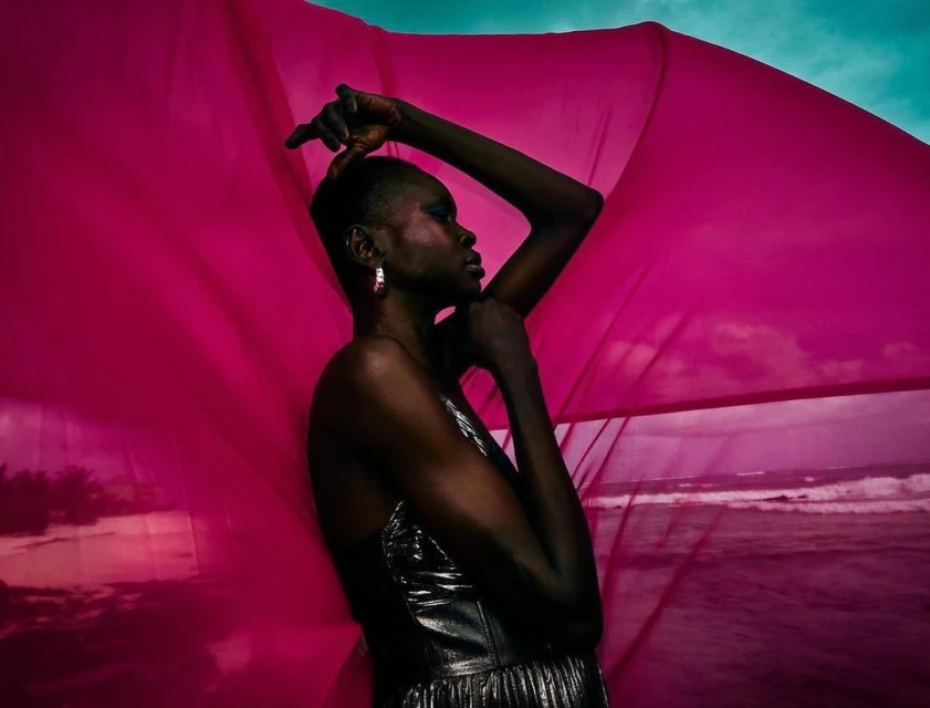 Woman in shiny dress poses with pink fabric against ocean backdrop in a striking fashion shot with dramatic lighting and styling.