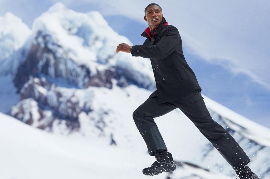 A man in a black coat and pants runs on a snowy surface with a snow-capped mountain range in the background, looking off to the right.