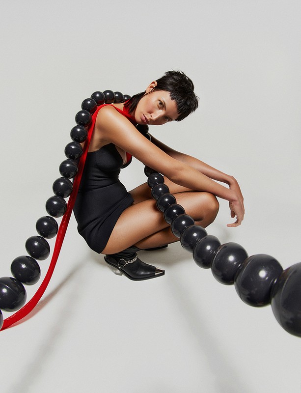 An adult woman in a black dress squats in a photo studio with a long strand of black spherical beads draped around her body.