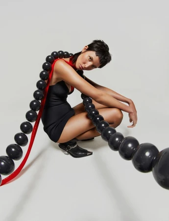 An adult woman in a black dress squats in a photo studio with a long strand of black spherical beads draped around her body.