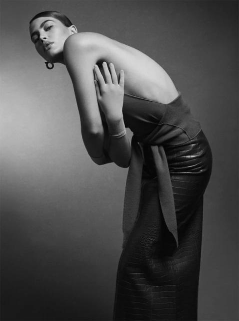 Monochrome studio shot of a woman in elegant dark dress, leaning forward with hands on her shoulders, looking at the camera.