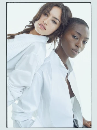Two models wearing white shirts posing together with heads leaning against each other against a white background