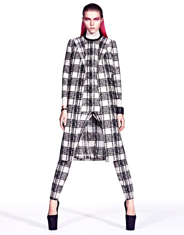 A model with pink hair poses in a black-and-white plaid coat and pants with platform heels in a studio setting, high fashion photography.
