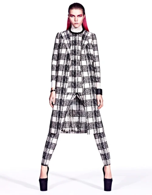 A model with pink hair poses in a black-and-white plaid coat and pants with platform heels in a studio setting, high fashion photography.