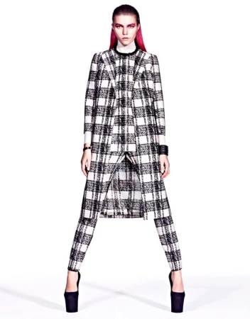 A model with pink hair poses in a black-and-white plaid coat and pants with platform heels in a studio setting, high fashion photography.