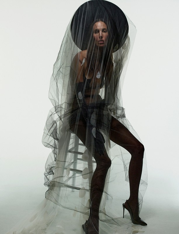 Model seated on a ladder wearing a sheer black veil and wide hat by photographer James Macari