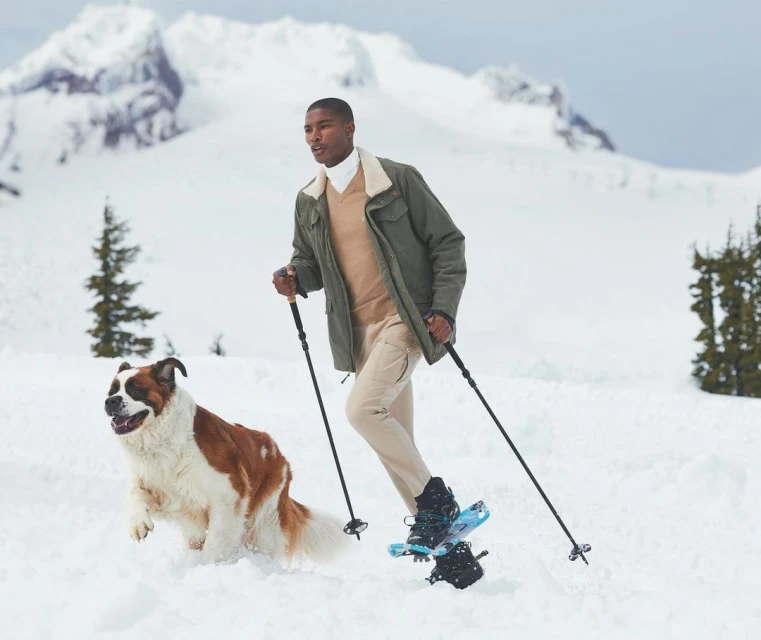 An African-American man snowshoeing with a Saint Bernard dog in a snowy mountain landscape with evergreen trees on a sunny day.