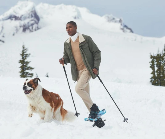 An African-American man snowshoeing with a Saint Bernard dog in a snowy mountain landscape with evergreen trees on a sunny day.