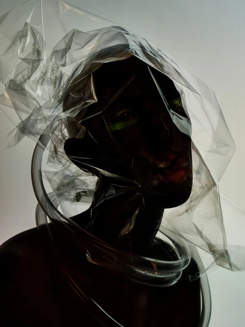 Person encased in transparent plastic and tubes against a light background. Dark skin and facial expression are partially visible.