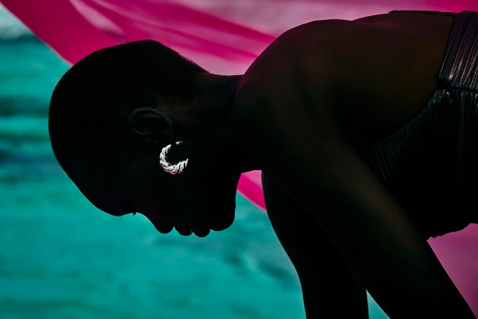 Silhouette of a Black model in a silver top and hoop earring with pink and teal fabric backdrop for fashion or beauty content.