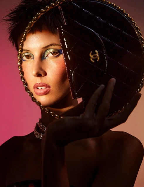 Close-up of an adult woman with creative makeup and a black quilted bag with a gold chain, posing against a warm pink gradient background.