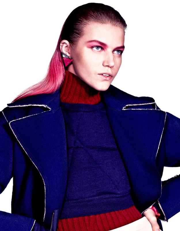 Fashionable adult female model with pink hair, bold pink eye makeup and a dark blue coat, posing in a studio with a white background.