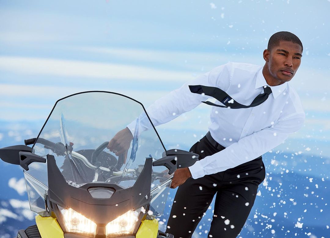 A man in a white dress shirt and black tie rides a yellow snowmobile across a snowy mountain landscape with a clear blue sky.