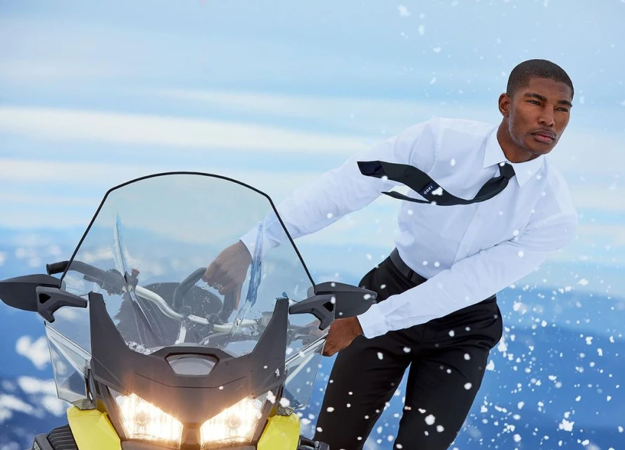 A man in a white dress shirt and black tie rides a yellow snowmobile across a snowy mountain landscape with a clear blue sky.