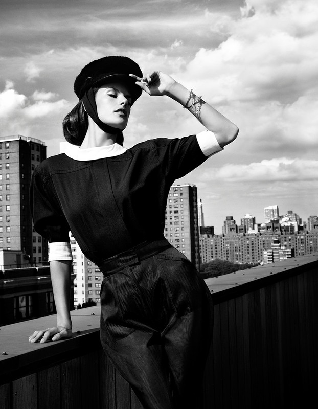 Monochrome fashion shot of a woman in vintage style clothing and cap, posing on a rooftop with a city skyline in the background.