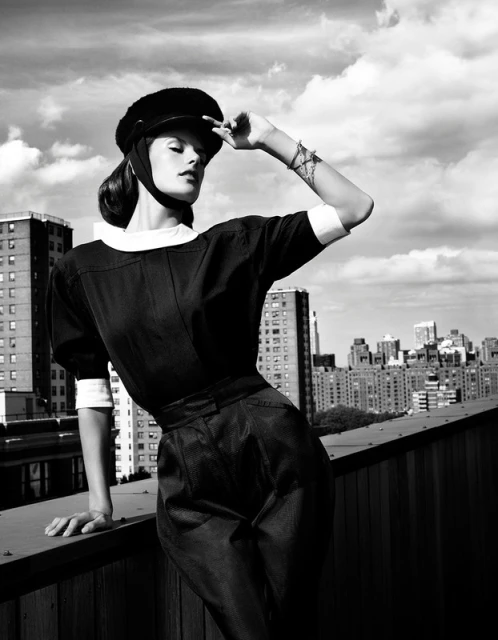 Monochrome fashion shot of a woman in vintage style clothing and cap, posing on a rooftop with a city skyline in the background.