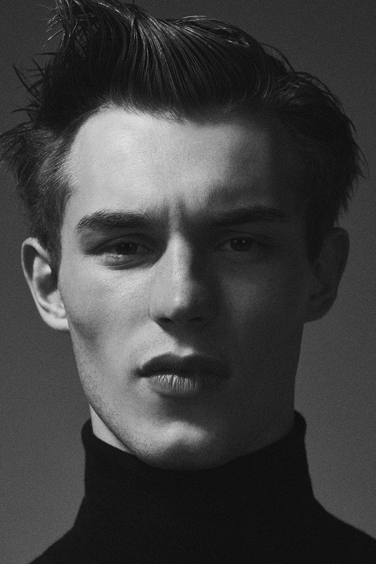 Monochrome headshot of a young adult male with dark hair styled upwards, wearing a black turtleneck sweater against a grey backdrop.